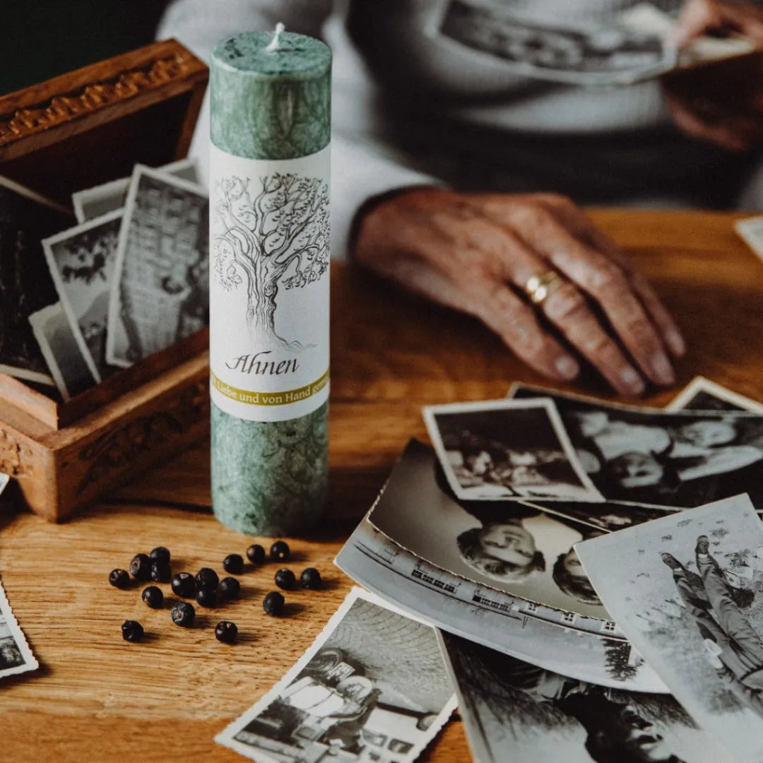 Dunkelgrüne Kräuterkerze "Ahnen" umgeben von alten Fotos & Wacholderbeeren, symbolisiert das Erbe der Vorfahren.