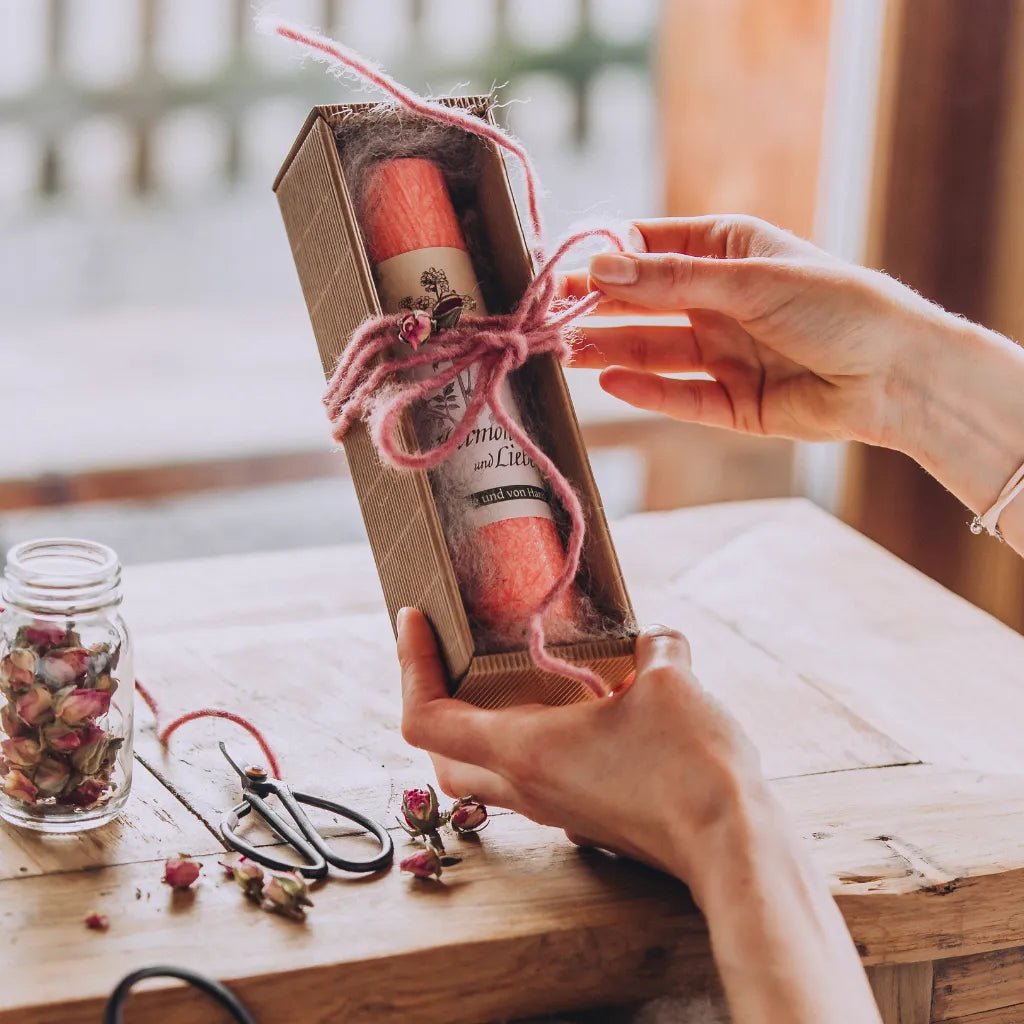 Hände binden eine rosa Schleife um eine Geschenkbox mit pinker Allgäuer Heilkräuterkerze.