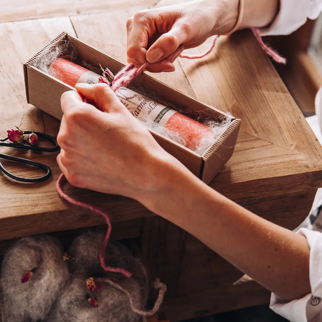 Hände binden eine rosa Schleife um eine Geschenkbox mit Allgäuer Heilkräuterkerze.