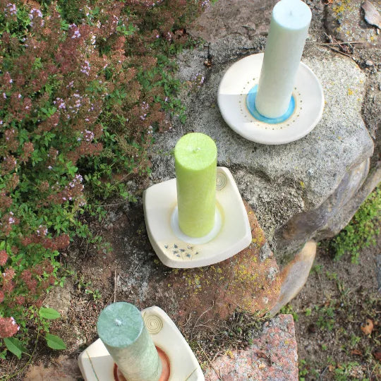 Allgäuer Heilkräuterkerzen auf handgetöpferten Kerzentellern mit Blüten- und Spiralverzierung im Garten.