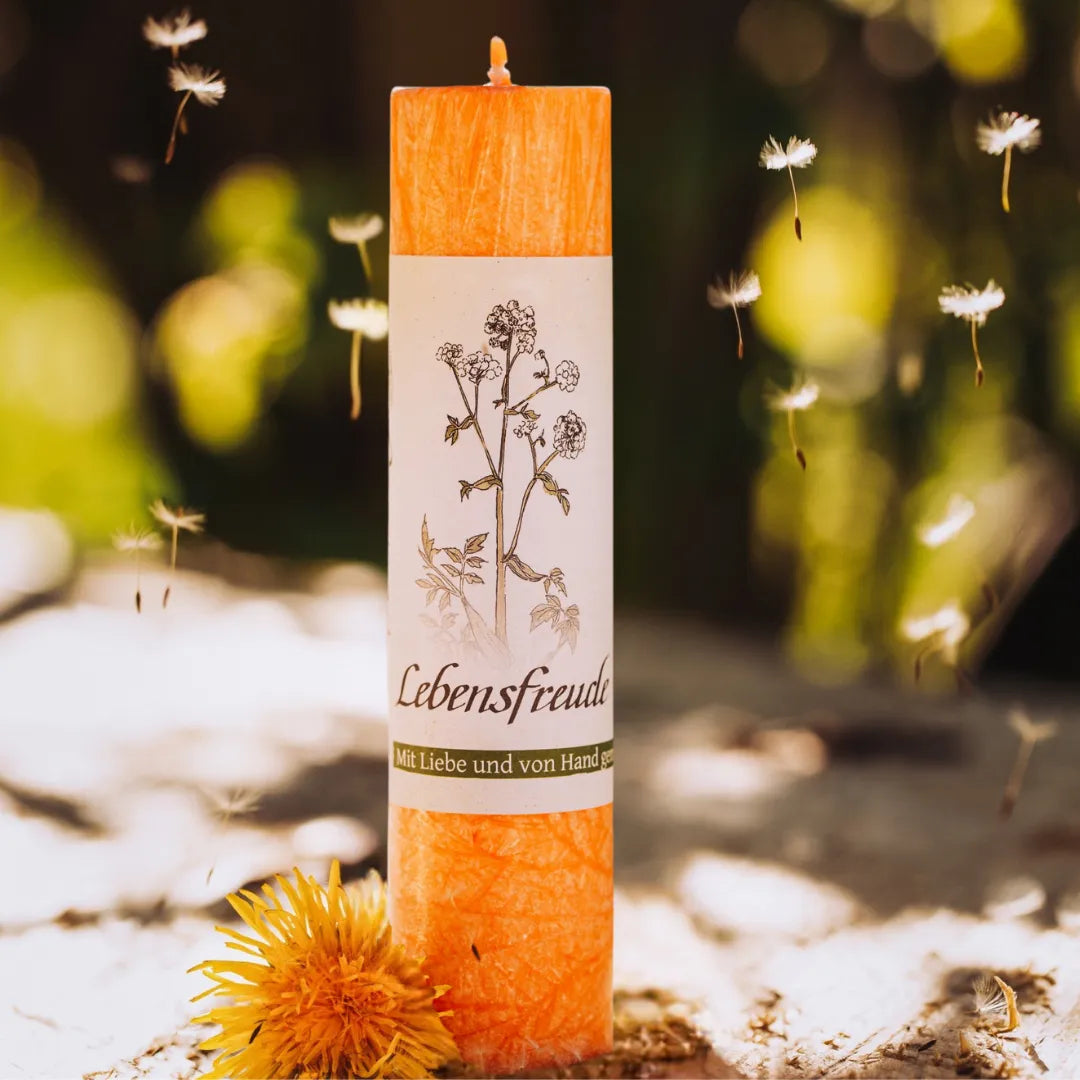 Orange Allgäuer Heilkräuterkerze "Lebensfreude" mit fliegenden Pusteblumen, für Frische und Freude.