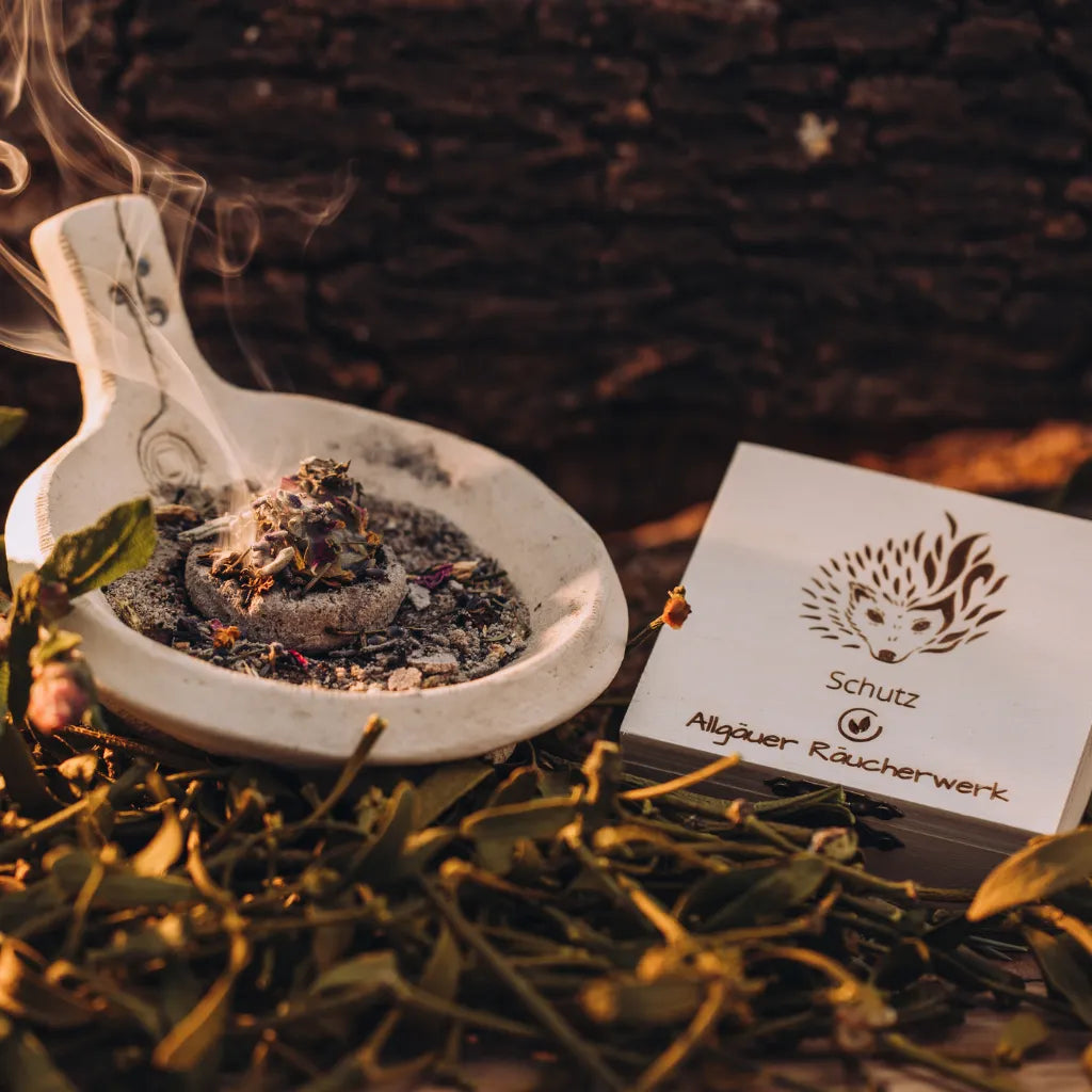 Stimmungsvolle Präsentation des Allgäuer Räucherwerks "Schutz" mit Igel-Box und rauchender Schale in natürlicher Umgebung.