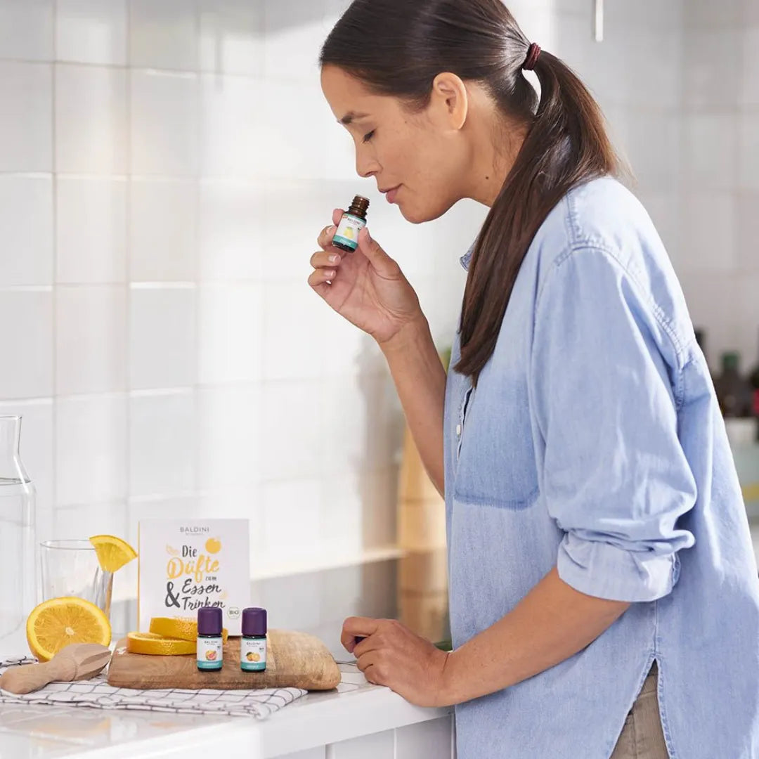 Frau riecht an Baldini Bio Zitronenöl in der Küche, mit weiteren Ölen und Rezeptbuch.