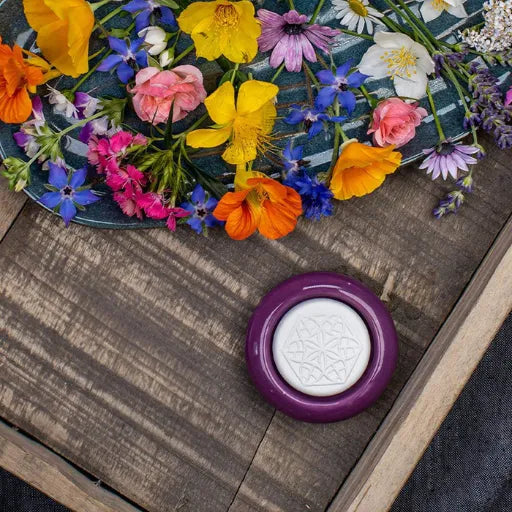 Baldini Duftstein in violetter Keramikfassung mit Lebensblume-Muster, umgeben von bunten Blüten.