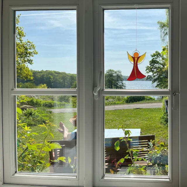 Sonnenfänger Engel in Rot-Gelb am Fenster mit Seeblick, vermittelt Gefühl von Schutz, Hoffnung und Geborgenheit.