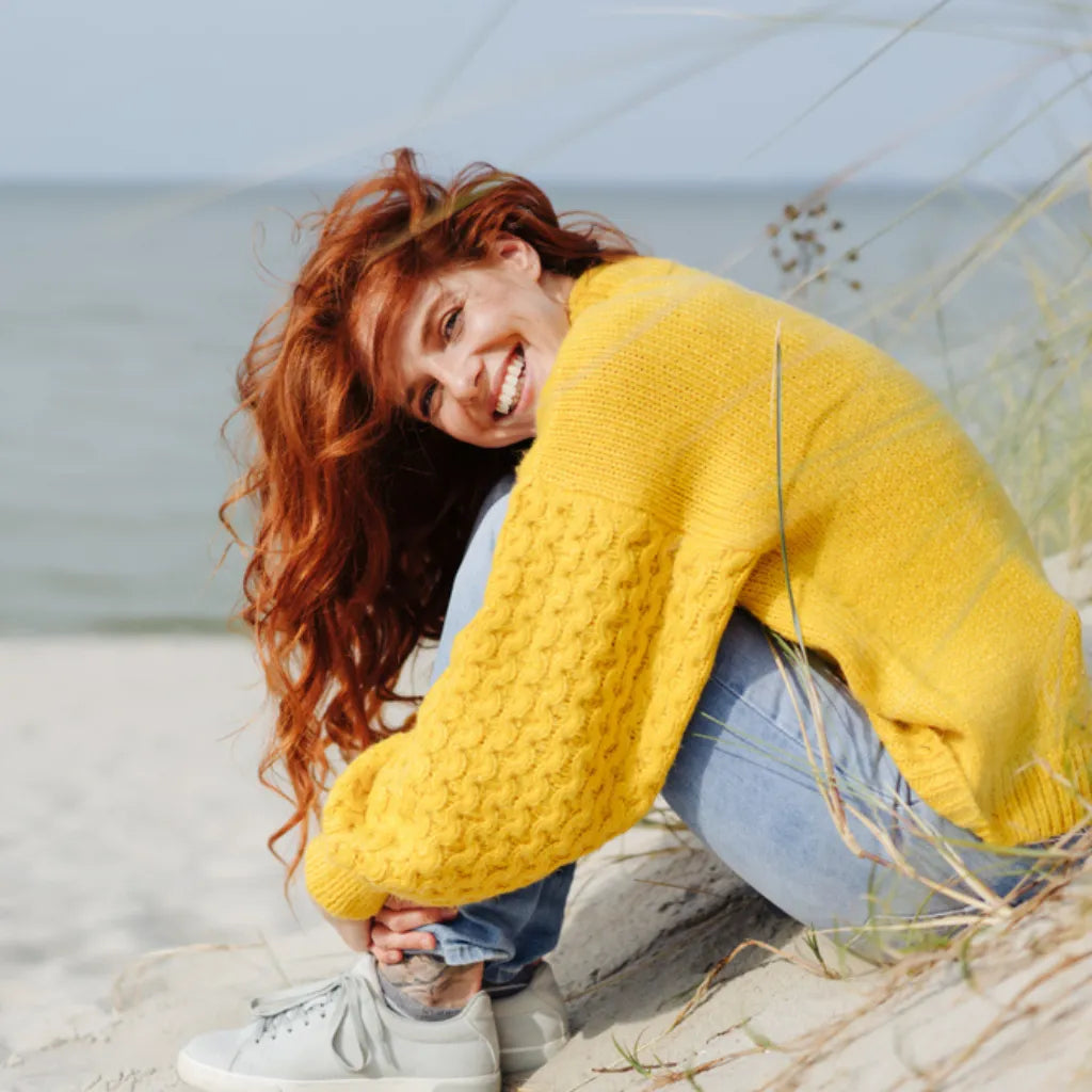 Frau lacht entspannt am Strand, symbolisiert innere Ruhe & Achtsamkeit durch Meditation im Hier und Jetzt.