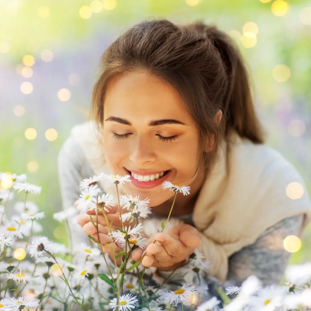 Junge Frau lächelt entspannt und riecht an Gänseblümchen auf einer sonnigen Wiese.