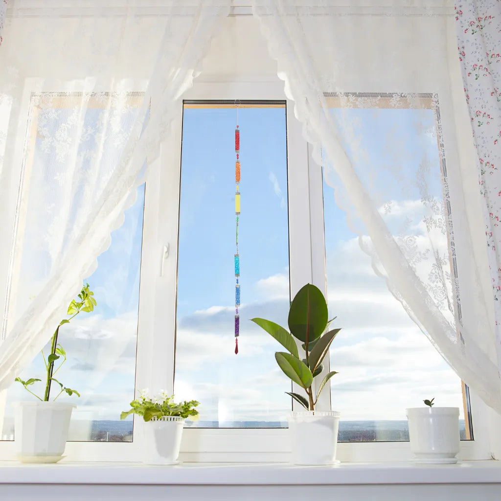 Die farbenfrohe Glaskette "Regenbogen" hängt als Fensterschmuck zwischen weißen Vorhängen und Topfpflanzen. Sie bringt lebendige Farben und ein schönes Lichtspiel in den Raum.