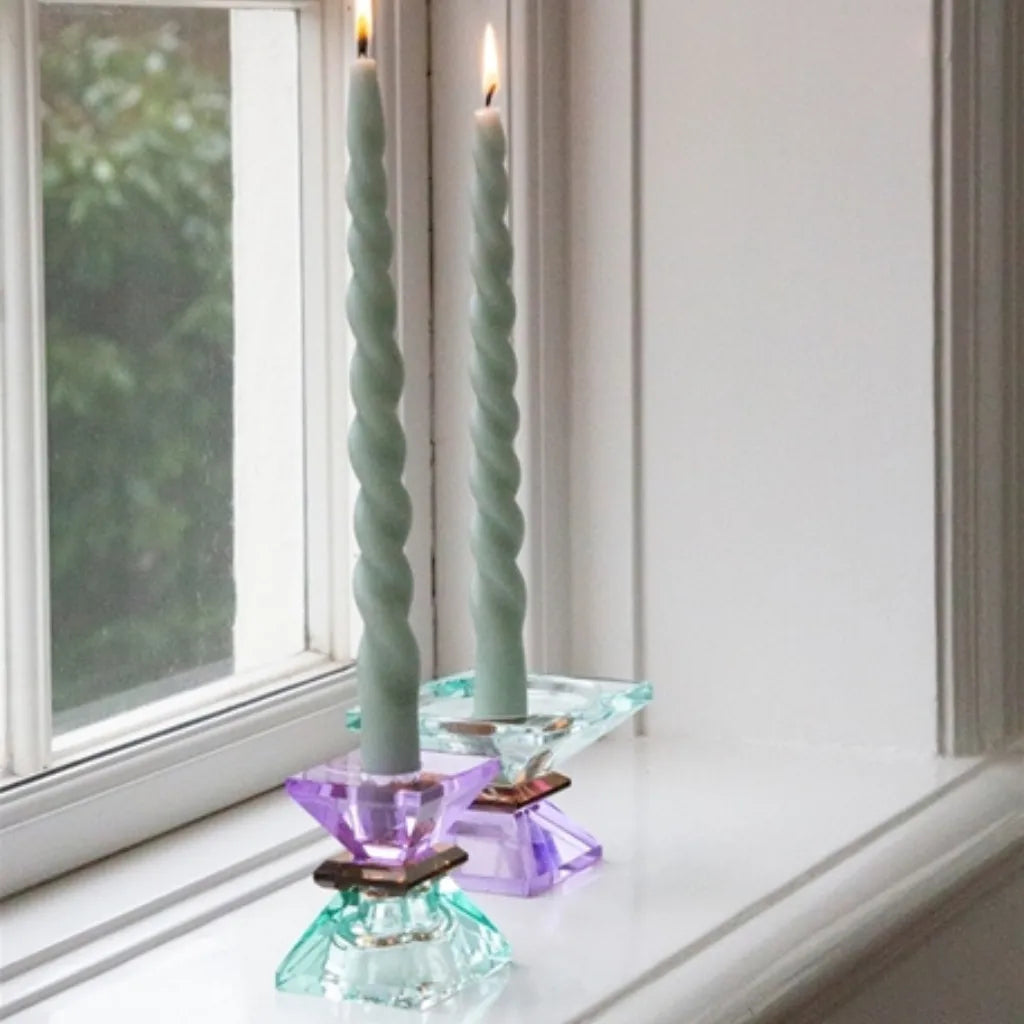 Zwei Kristall-Kerzenhalter in hellminze/dunkelbraun/violett mit brennenden grünen Stabkerzen auf einer Fensterbank.