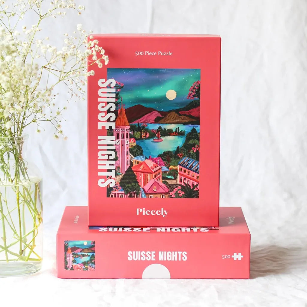 Zwei Piecely Puzzle 500 "Suisse Nights" Boxen, eine liegend, eine stehend, arrangiert mit zarten weißen Blumen. Zeigt die ästhetische Präsentation des Puzzles.