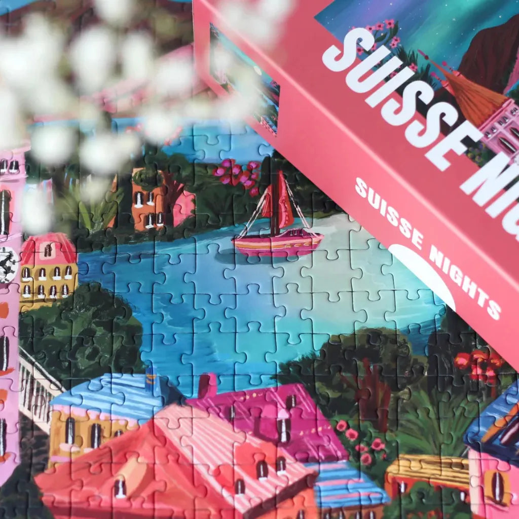 Nahaufnahme des teilweise gelegten Piecely Puzzles 500 "Suisse Nights" mit der Verpackung im Hintergrund. Fokussiert auf die bunten Häuser, das Segelboot und den See.