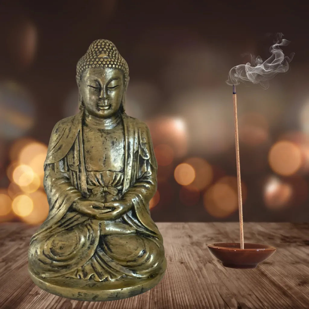 Ein brennendes Räucherstäbchen in einem braunen Keramikhalter, platziert neben einer sitzenden Buddha-Statue vor einem verschwommenen, warmen Hintergrund, symbolisieren Meditation und Entspannung.
