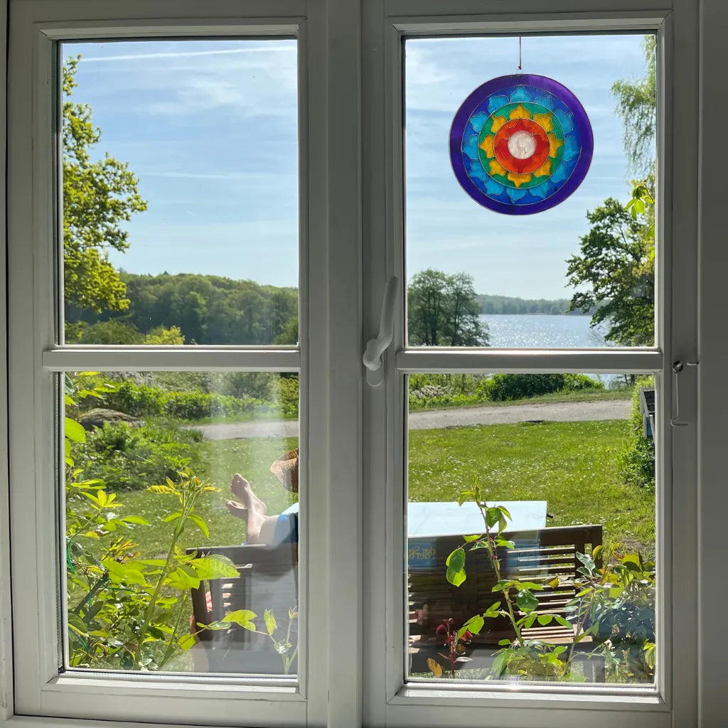 Der bunte Sonnenfänger "Lotus Chakra Regenbogen" hängt an einem Fenster und fängt das Licht ein. Er bietet einen harmonischen Ausblick auf einen See und die grüne Landschaft, ideal für meditative Momente.