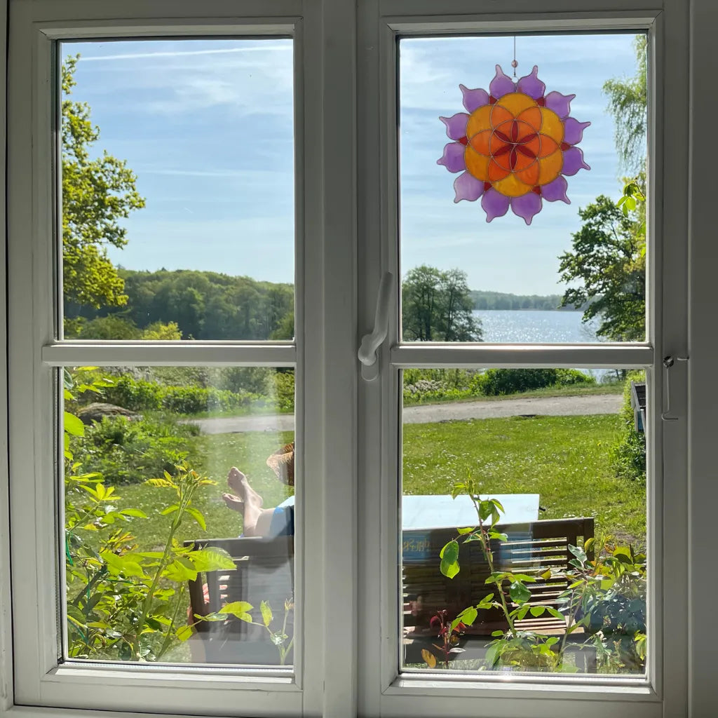 Der Sonnenfänger "Lotus Saat des Lebens" aus farbigem Glas hängt in einem Fenster. Er bietet einen Blick auf einen See und die grüne Landschaft und sorgt für eine harmonische und spirituelle Atmosphäre.