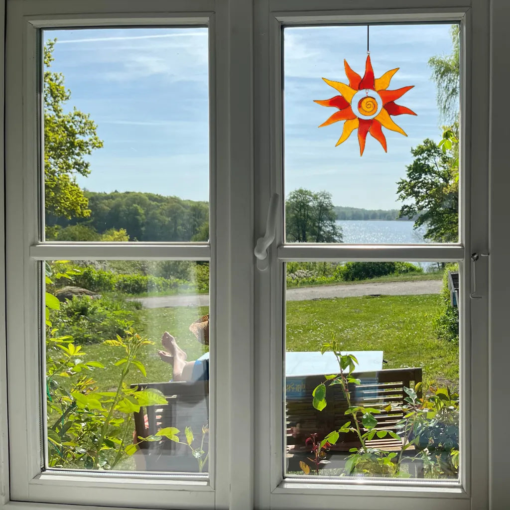 Der orange-gelbe Sonnenfänger Sonne aus Glas (15cm) hängt dekorativ an einem Fenster. Er fängt das Licht ein und bietet eine malerische Aussicht auf einen See und die grüne Landschaft im Hintergrund.