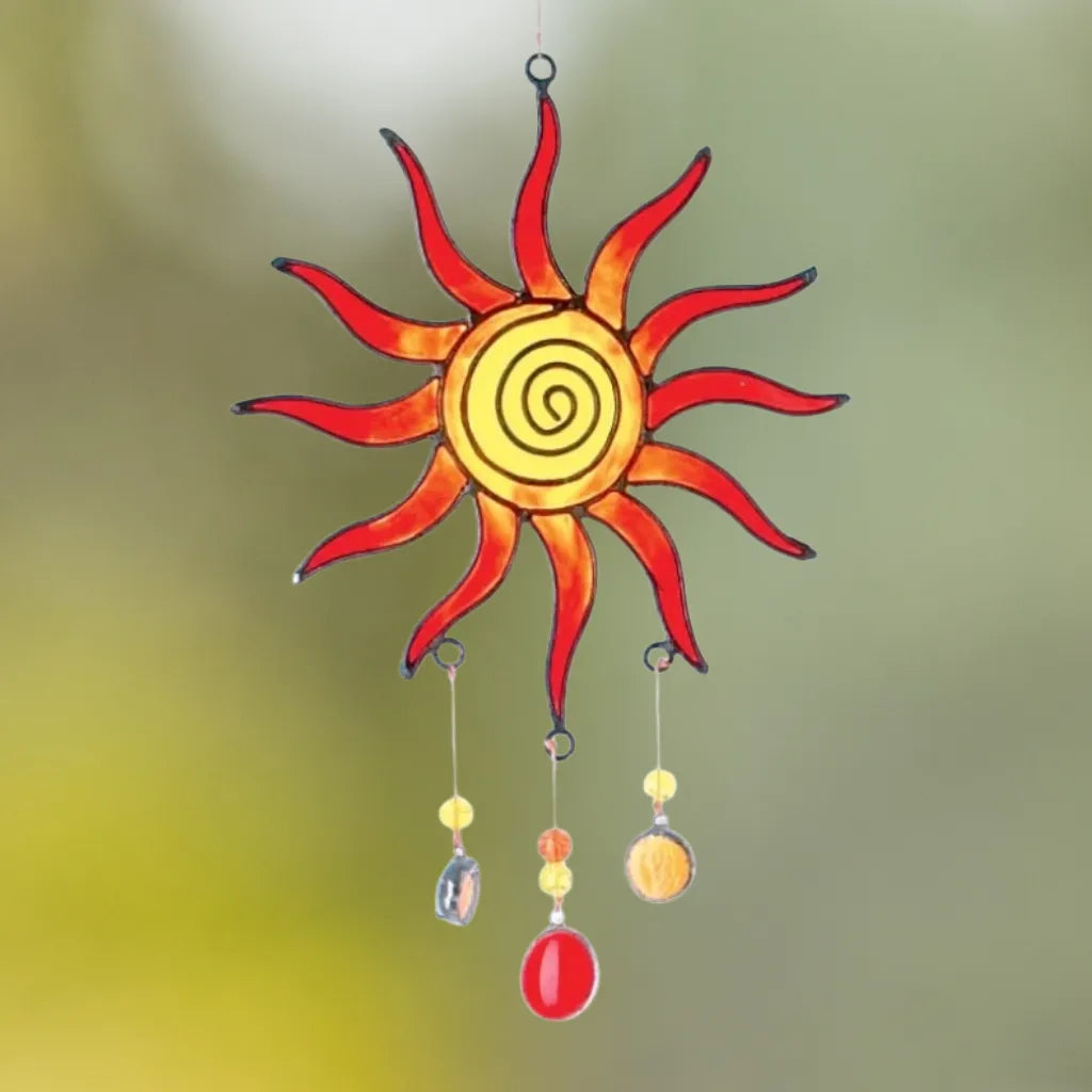 Glänzender Sonnenfänger "Sonne Spirale" aus mehrfarbigem Glas (Rot-Gelb-Orange), mit hängenden Perlen, vor einem unscharfen grünen und gelben Hintergrund. Erzeugt ein lebendiges Lichtspiel.