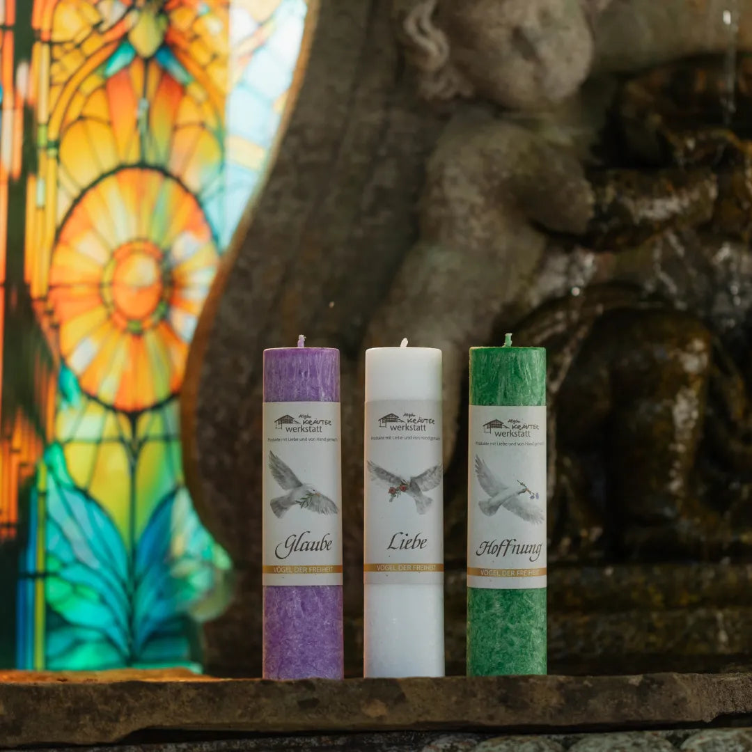 Drei Kerzen (lila, weiß, grün) mit Etiketten "Glaube", "Liebe", "Hoffnung" und Taubenmotiv, stehend vor einem farbigen Kirchenfenster und einer steinernen Figur.