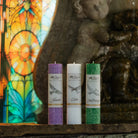 Drei Kerzen (lila, weiß, grün) mit Etiketten "Glaube", "Liebe", "Hoffnung" und Taubenmotiv, stehend vor einem farbigen Kirchenfenster und einer steinernen Figur.