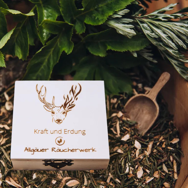 Holzbox "Allgäuer Räucherwerk Kraft und Erdung" mit Hirsch-Motiv auf einem Bett aus Kräutern.