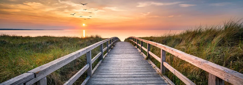 Holzsteg führt über Dünen zum ruhigen Meer bei Sonnenuntergang, ideal für Meditation und Entspannung.