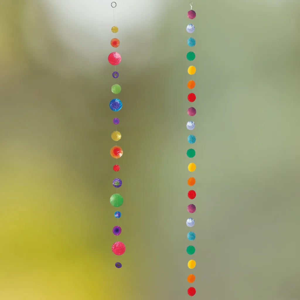 Zwei bunte Capiz-Muschelketten, Variante 1 und Variante 2, hängen vor einem weichen, unscharfen Hintergrund und erzeugen ein leichtes, maritimes Ambiente als Fenster- oder Raumdeko.