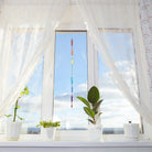 Die farbenfrohe Glaskette "Regenbogen" hängt als Fensterschmuck zwischen weißen Vorhängen und Topfpflanzen. Sie bringt lebendige Farben und ein schönes Lichtspiel in den Raum.