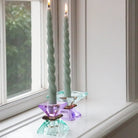 Zwei Kristall-Kerzenhalter in hellminze/dunkelbraun/violett mit brennenden grünen Stabkerzen auf einer Fensterbank.