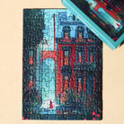 Das fertig gelegte 99 Teile Piecely Minipuzzle "Midnight Walk" neben der dazu passenden, türkisfarbenen Verpackung. Eine atmosphärische Ansicht der nächtlichen Stadt mit Gebäuden und Grün.