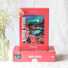 Zwei Piecely Puzzle 500 "Suisse Nights" Boxen, eine liegend, eine stehend, arrangiert mit zarten weißen Blumen. Zeigt die ästhetische Präsentation des Puzzles.