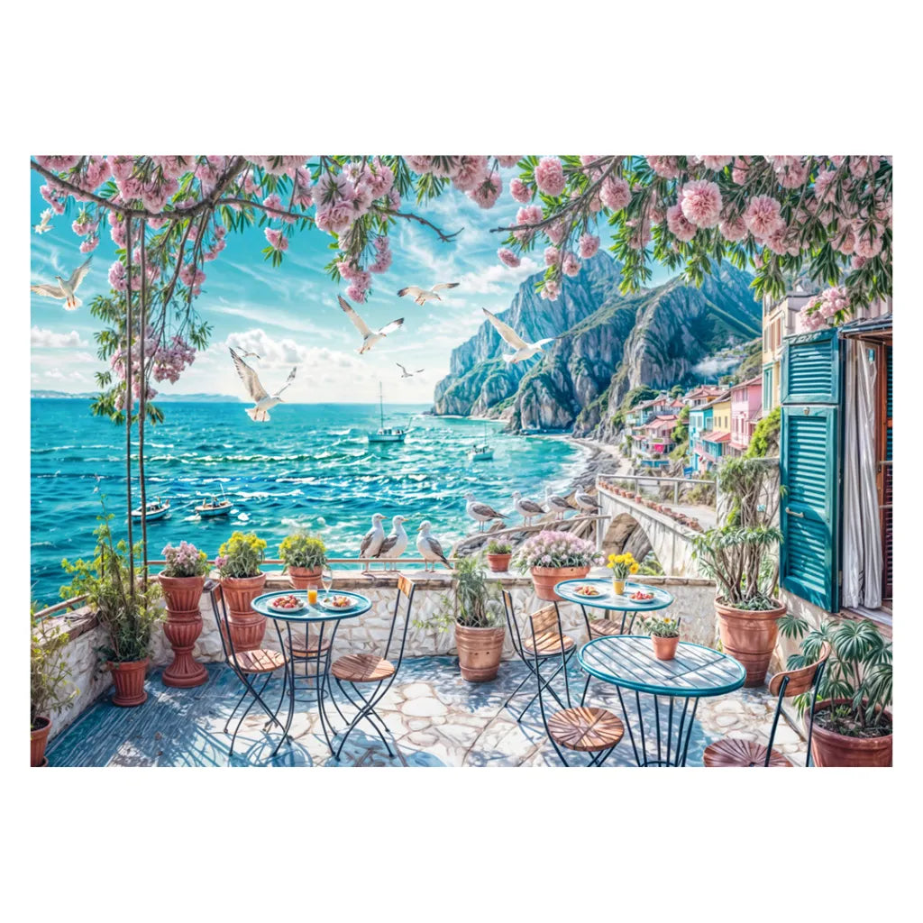 Detailliertes Motiv des Schmidt Puzzles 1000 Teile "Terrasse am Meer". Ein sonniger Balkon mit Tischen, blühenden Pflanzen und Blick auf das blaue Mittelmeer und Küstenstadt.