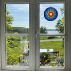 Der bunte Sonnenfänger "Lotus Chakra Regenbogen" hängt an einem Fenster und fängt das Licht ein. Er bietet einen harmonischen Ausblick auf einen See und die grüne Landschaft, ideal für meditative Momente.