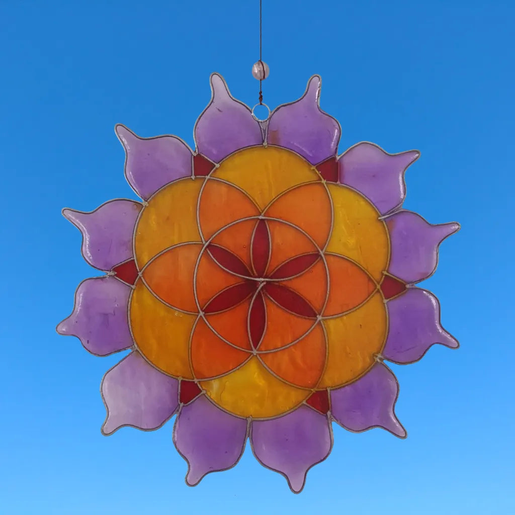 Der Sonnenfänger "Lotus Saat des Lebens" aus farbigem Glas (Lila, Orange, Gelb) hängt vor einem strahlend blauen Himmel. Er fängt das Licht ein und erzeugt ein wunderschönes Farbenspiel.