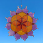 Der Sonnenfänger "Lotus Saat des Lebens" aus farbigem Glas (Lila, Orange, Gelb) hängt vor einem strahlend blauen Himmel. Er fängt das Licht ein und erzeugt ein wunderschönes Farbenspiel.