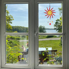 Der Sonnenfänger "Sonne Spirale" hängt in einem Fenster mit Blick auf einen See und grüne Landschaft. Das rote und gelbe Glasobjekt mit Perlen fängt das Sonnenlicht wunderschön ein.